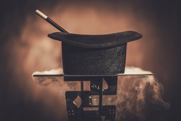 A black top hat sits upsidown on a smoking table, held up by metal playing cards.  A magic wand is inside the hat